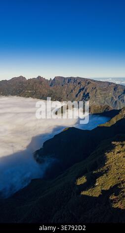 Aus der Vogelperspektive auf die zentralen Berge Madeiras in der Nähe von Pico do Arieiro, während Wolken Täler füllen. Das Licht am frühen Morgen wirft lange Schatten auf zerklüftete grüne Hänge und Stockfoto