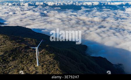 Aus der Vogelperspektive über Madeira zeigt eine einsame Windturbine auf einem Bergrücken über einem Wolkenmeer, mit frühem oder spätem Licht, das kühle Schatten auf tiefem Grau wirft Stockfoto