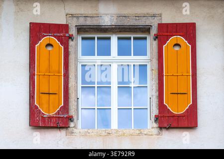 Rustikale rot-orange Fensterläden mit weißer Zierleiste betonen ein klassisches europäisches Fenster vor einer strukturierten Gipsmauer in Porrentruy, Schweiz. Stockfoto