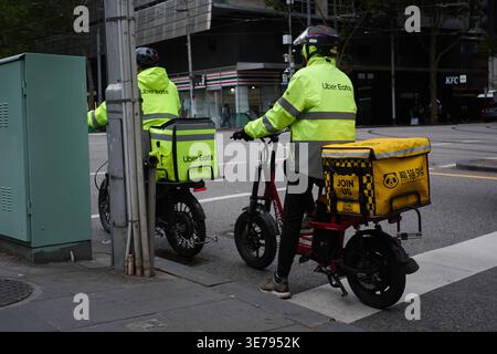 Zwei Essenskuriere auf Fahrrädern, beide tragen Ausrüstung der Marke Uber Eats, sitzen an einer Kreuzung in Melbourne Stockfoto