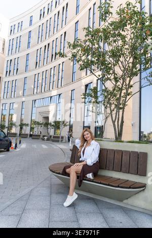 Eine Frau sitzt auf einer runden Bank in einem modernen Geschäftsviertel und genießt eine friedliche Mittagspause. Ihre entspannte Haltung und ihr stilvolles Outfit spiegeln urbanes Ambiente wider Stockfoto