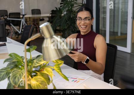Weibliche Arbeiterin in kastanienbraunem Oberteil lächelnd, Smartphone am modernen Schreibtisch mit Lampe und Karten Stockfoto