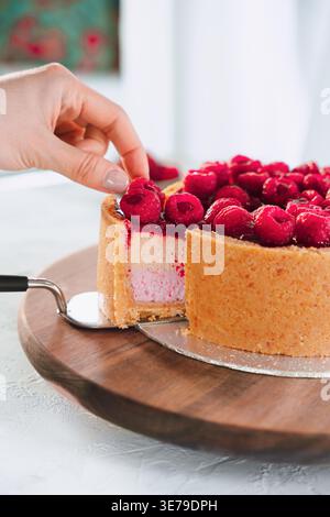 Hand nimmt ein Stück Himbeerkuchen aus Holzbrett mit frischen Beeren in der hellen Küche, Dessert-Moment, hausgemachte Backen Stockfoto