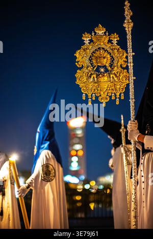 Ein Nazarener aus der Bruderschaft La Estrella hält ein Banner während einer Karwoche bei Nacht auf der Triana-Brücke in Sevilla. Wolkenkratzer Torre Sevilla Stockfoto