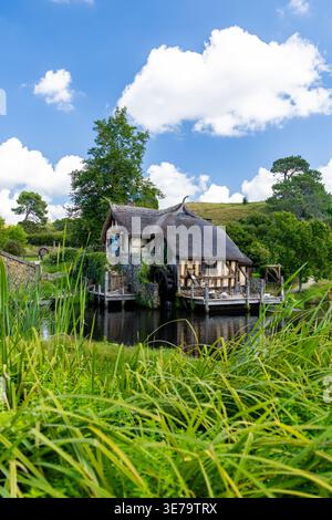 Blick auf ein malerisches, strohgedecktes Cottage auf einem ruhigen Teich inmitten von lebhaftem Grün unter hellem Himmel, Hobbiton Movie Set, Waikato Region, New Ze Stockfoto