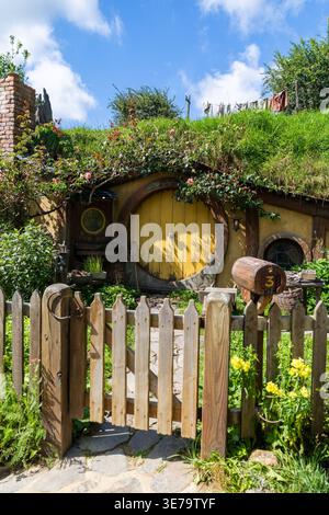 Blick auf ein malerisches Hobbit-Loch mit einer lebendigen gelben Tür, eingebettet in einen grasbewachsenen Hügel, eingerahmt von einem rustikalen Holzzaun, Hobbiton Movie Set, Waikato Regio Stockfoto