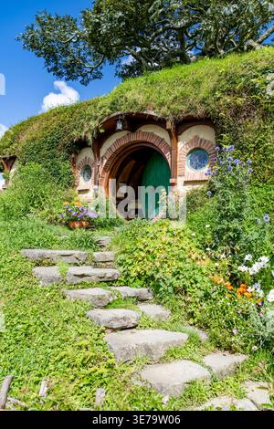 Blick auf ein malerisches Hobbit-Loch, eingebettet in einen grünen Hügel mit einer runden grünen Tür und Steintreppen im Hobbiton Movie Set, Matamata, Neuseeland. Stockfoto