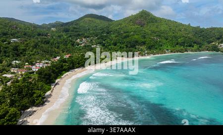 Klare türkisfarbene Wellen ziehen über große, glatte Felsen am weißen Sandstrand, umgeben von Grün. Seychellen, Mahe. Baie Lazare. Stockfoto