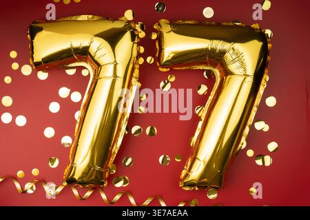 77. Geburtstag. Goldene festliche Ballons Zahl 77-jähriges Jubiläum auf rotem Hintergrund mit Konfetti. Geburtstag oder Hochzeit. Stockfoto