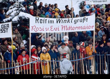 Zuschauer versammeln sich bei den XII. Olympischen Winterspielen in Innsbruck, um die Athleten anzufeuern. Im Vordergrund ist ein Banner zu sehen, das die Skirennläuferinnen Rosi und Evi Mittermaier aus Reit im Winkl grüßt. Die Szene zeigt die winterliche Atmosphäre mit Schnee und Bäumen. Österreich, 1976 Stockfoto