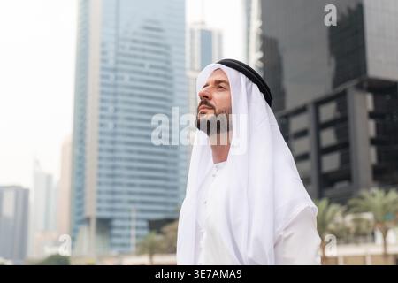 Arabischer Geschäftsmann in einem traditionellen weißen Kandura steht in Dubai Marina mit Blick auf die Skyline. Stockfoto