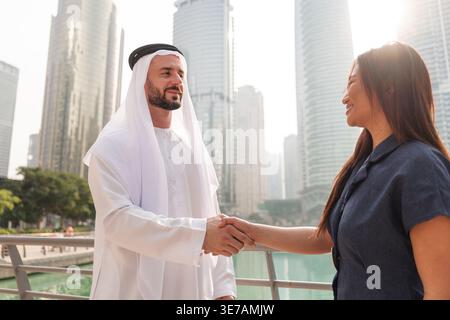 Eine asiatische Frau und ein Emirati schütteln in Dubai Marina die Hände, während sie über einen Immobilieninvestitionsvertrag in der Gegend diskutieren. Stockfoto