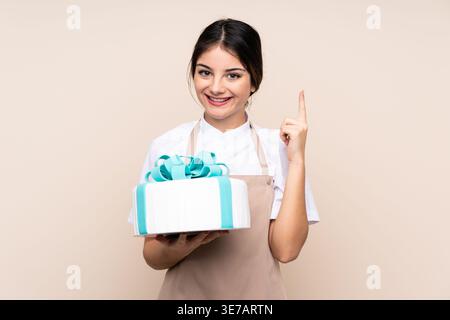 Hispanische Konditorin, die im Studio vor beigefarbenem Hintergrund Kuchen hält Stockfoto