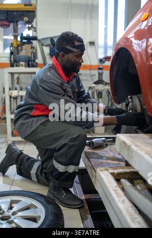 Ernster schwarzer Mechaniker kniete sich nieder und trug Handschuhe, die das Autorad in der Werkstatt wechseln Stockfoto