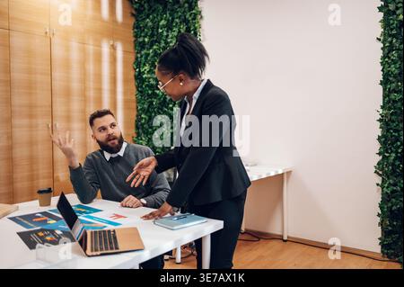 Schwarze Geschäftsfrau in Brille steht und diskutiert Projekte mit chLifestyles auf Tisch mit Laptop während eines Meetings mit männlichem Kollegen im Büro Stockfoto