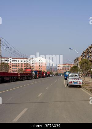 Guangshui City, China - 22. Februar 2026: Eine breite Stadtstraße mit mehreren schweren Lkw, die an der Seite geparkt sind. Im Hintergrund ein industrielles Werk Stockfoto