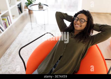 Schwarze Frau mit lockigen Haaren, die Brille trägt, sitzt mit Händen hinter dem Kopf und geschlossenen Augen auf Sitzsack im Wohnzimmer zu Hause Stockfoto