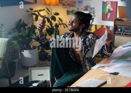 Seitenansicht eines lächelnden Indianers, der auf einem Stuhl sitzt und Selfie auf dem Smartphone im Studio macht Stockfoto