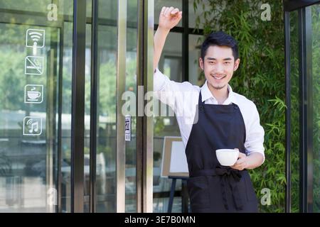 Ein chinesischer Mann mit Schürze, der eine Kaffeetasse hält und sich an die Tür des Cafés lehnt Stockfoto