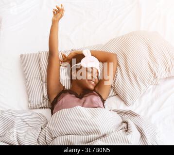 Schwarze Frau mit Augenmaske auf der Stirn, die Arme nach dem Aufwachen beim Liegen auf dem Bett zu Hause dehnt Stockfoto