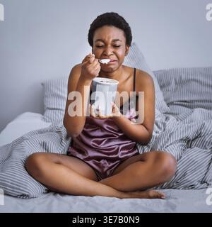 Weinende Schwarze Frau mit kurzen Haaren isst Eis aus dem Eimer, während sie zu Hause mit überkreuzten Beinen auf dem Bett sitzt Stockfoto