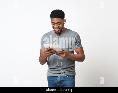 Lächelnder Schwarzer Mann, der ein Tablet mit Ohrhörern benutzt, während er im Studio vor weißem Hintergrund steht Stockfoto