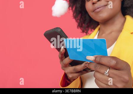 Hispanische Frau mit Afro trägt gelbe Jacke und Weihnachtsmann-Hut, die Kreditkarte hält und online Smartphone-Shopping nutzt, während sie gegen Rosa steht... Stockfoto