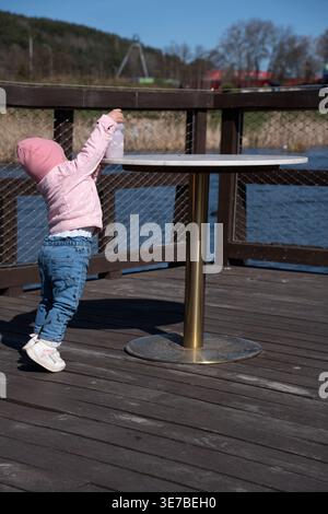 Kind in rosafarbenem Hut und Jacke, das auf einem runden Kaffeetisch auf einer hölzernen Uferterrasse unter einem klaren blauen Himmel mit weit entfernten Bäumen, weich, nach einer Flasche greift Stockfoto