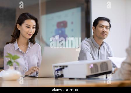 Chinesische Geschäftsfrau, die am Laptop arbeitet, sitzt neben einem männlichen Kollegen im Konferenzraum Stockfoto