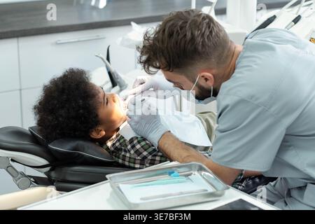 Schwarzer Junge, der mit kaukasischem männlichen Zahnarzt behandelt wird, während er auf einer Zahnarztklinik sitzt Stockfoto