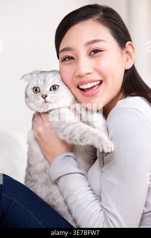 Lächelnde Chinesin, die schottische Faltkatze trägt und umarmt, während sie zu Hause auf dem Sofa sitzt Stockfoto