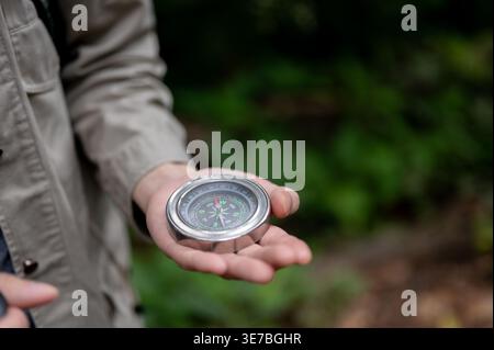Asiatischer männlicher Wanderer mit brauner Jacke und Blick auf den Kompass, während er im Wald steht Stockfoto