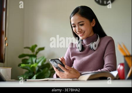 Lächelnde asiatische Frau mit Kopfhörern am Hals, die ihr Smartphone benutzt, während sie zu Hause mit Büchern am Tisch sitzt Stockfoto