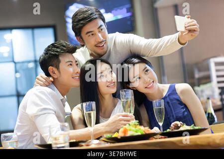 Ein lächelnder Pazifikinsel-Mann macht Selfie auf dem Smartphone mit chinesischen Freunden, die zusammen im Restaurant zu Abend essen Stockfoto