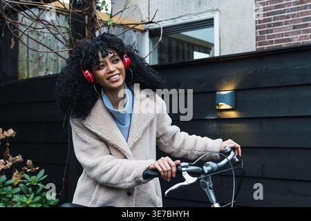 Lächelnde Mixed Race Frau mit lockigem Haar, die warme Jacke und Kopfhörer trägt und wegblickt, während sie auf der Straße Fahrrad fährt Stockfoto