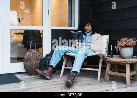 Gemischte Rassenfrau mit lockigen Haaren, die warme Jacke trägt, die Buch liest, während sie zu Hause auf dem Stuhl auf der Veranda sitzt Stockfoto