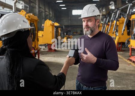 Schwarze Journalistin, die einen Sicherheitshelm mit Mikrofon trägt und einen kaukasischen Mann befragt, der tagsüber im Lagerhaus steht Stockfoto