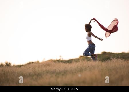 Schwarze Frau mit lockigem Afro, die einen roten Schal hält, läuft bei Sonnenaufgang auf der Wiese Stockfoto