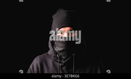 Hispanischer Mann mit schwarzem Hoodie, Mütze und Maske, die auf schwarzem Hintergrund stehen Stockfoto