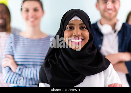 Fröhliche schwarze muslimische Geschäftsfrau im Hijab, die mit überkreuzten Armen neben männlichen und weiblichen Freunden im Studio an der Wand steht Stockfoto
