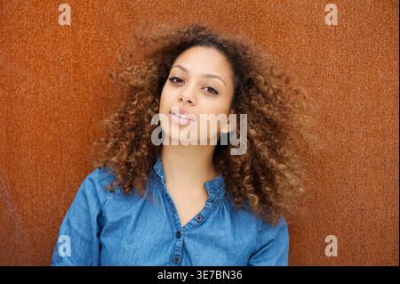 Lächelnde Mixed Race Frau mit lockigen Haaren, die blaue lange Ärmel trägt und an braunen Wänden steht Stockfoto