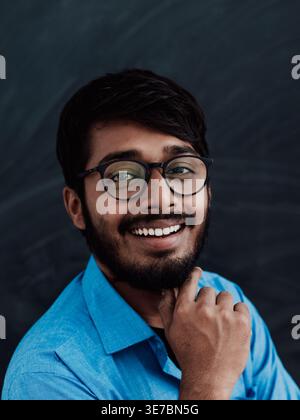 Lächelnder indischer Mann in blauen Langarmen mit Brille, die mit Hand am Kinn an der Tafel im Klassenzimmer steht Stockfoto