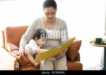 Chinesische Großmutter und kleines Mädchen lesen Buch, während sie zu Hause zusammen auf dem Sofa sitzen Stockfoto