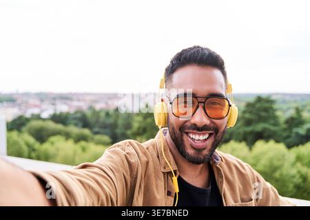 Hispanischer Mann mit Bart, Sonnenbrille und Kopfhörer, der tagsüber Selfie macht, während er draußen steht Stockfoto