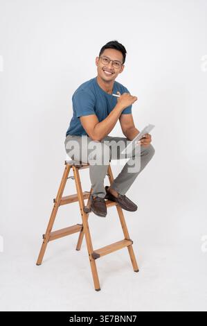 Lächelnder asiatischer Mann mit Stift, der Tablette hält, sitzt auf hölzerner Leiter vor weißem Hintergrund im Studio Stockfoto