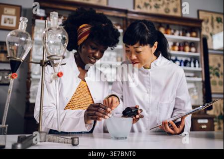 Schwarze Chemikerin gießt Zutat in Mörtel, wie japanische Kollegin zeigt und beobachtet stehende Holding Chart in Vintage-Apotheke Stockfoto