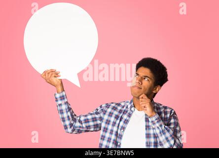 Verwirrter Schwarzer Mann mit der Hand in der Nähe des Gesichts, der eine leere Sprechblase hält, die im Studio vor rosa Hintergrund steht Stockfoto