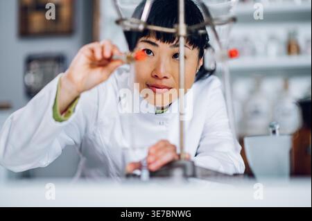 Japanische Chemikerin im Laborkittel, die sich auf das Gegenöffnungsventil des Trenntrichters stützt und es in das Becherglas einer Vintage-Apotheke fließen lässt Stockfoto