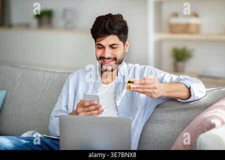 Lächelnder arabischer Mann mit Bart, der Smartphone benutzt und Kreditkarte hält, sitzend auf Sofa mit Laptop im Wohnzimmer zu Hause Stockfoto