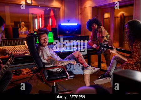 Eine hispanische Produzentin mit Afro sitzt in der Nähe eines männlichen Managers, der dem Gitarristen und Sänger im Musikstudio zuhört Stockfoto
